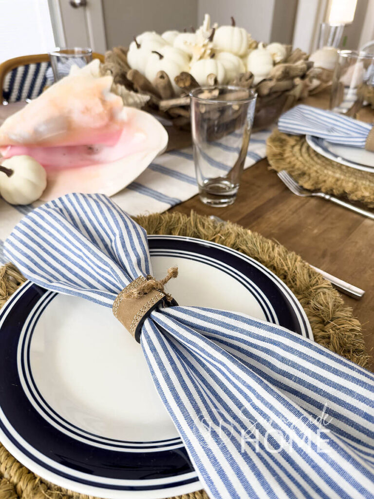 Coastal and fall blue Thanksgiving table decor with a blue and white table runner, and a centerpiece featuring white mini pumpkins with starfish, and scattered seashells and mini pumpkins. The placesettings feature a round straw placemat, blue rimmed dinner plate, light blue stripped napkin in a DIY leather belt napkin ring.