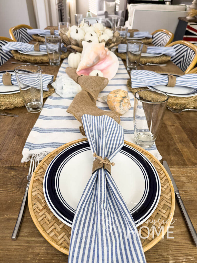 Coastal and fall blue Thanksgiving table decor with a blue and white table runner, and a centerpiece featuring white mini pumpkins with starfish, and scattered seashells and mini pumpkins. The placesettings feature a round woven grass placemat, blue rimmed dinner plate, light blue stripped napkin in a DIY leather belt napkin ring.
