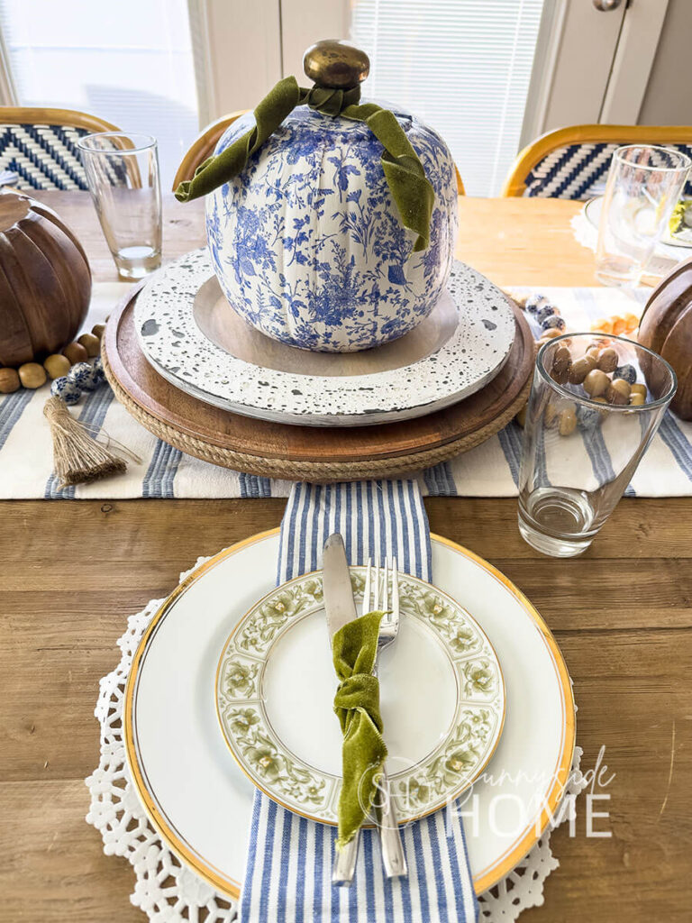 Elegant fall blue Thanksgiving table decor with a blue and white table runner topped with a round wooden tray and charger topped with a blue and white DIY chinoiserie pumpkin with a brass door knob stem and embellished with green velvet ribbon. The placesetting features a white doily placemat, gold rimmed thrift store plates, blue and white stripped napkins topped with a green and white salad place and the flatware it tied with a green velvet ribbon.