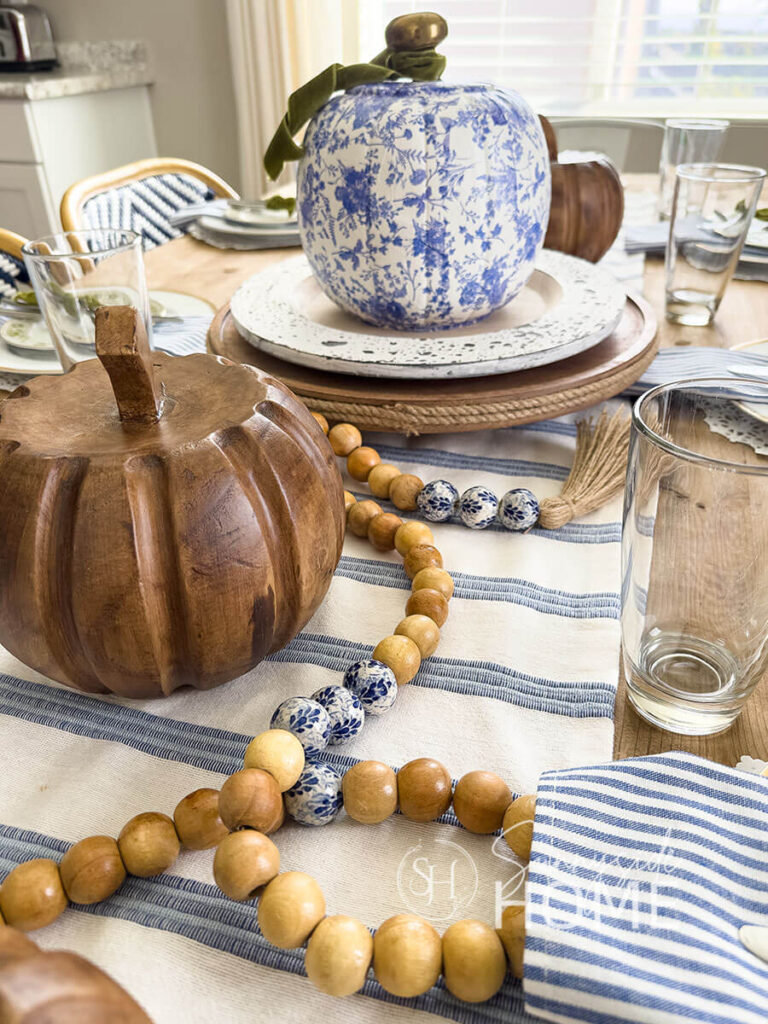Elegant fall blue Thanksgiving table decor with a blue and white table runner topped with a round wooden tray and charger topped with a blue and white DIY chinoiserie pumpkin with a brass door knob stem and embellished with green velvet ribbon, scattered along the table runner are wood look pumpkins and a wood and chinoiserie bead garland. The placesetting features a white doily placemat, gold rimmed thrift store plates, blue and white stripped napkins topped with a green and white salad place and the flatware it tied with a green velvet ribbon.