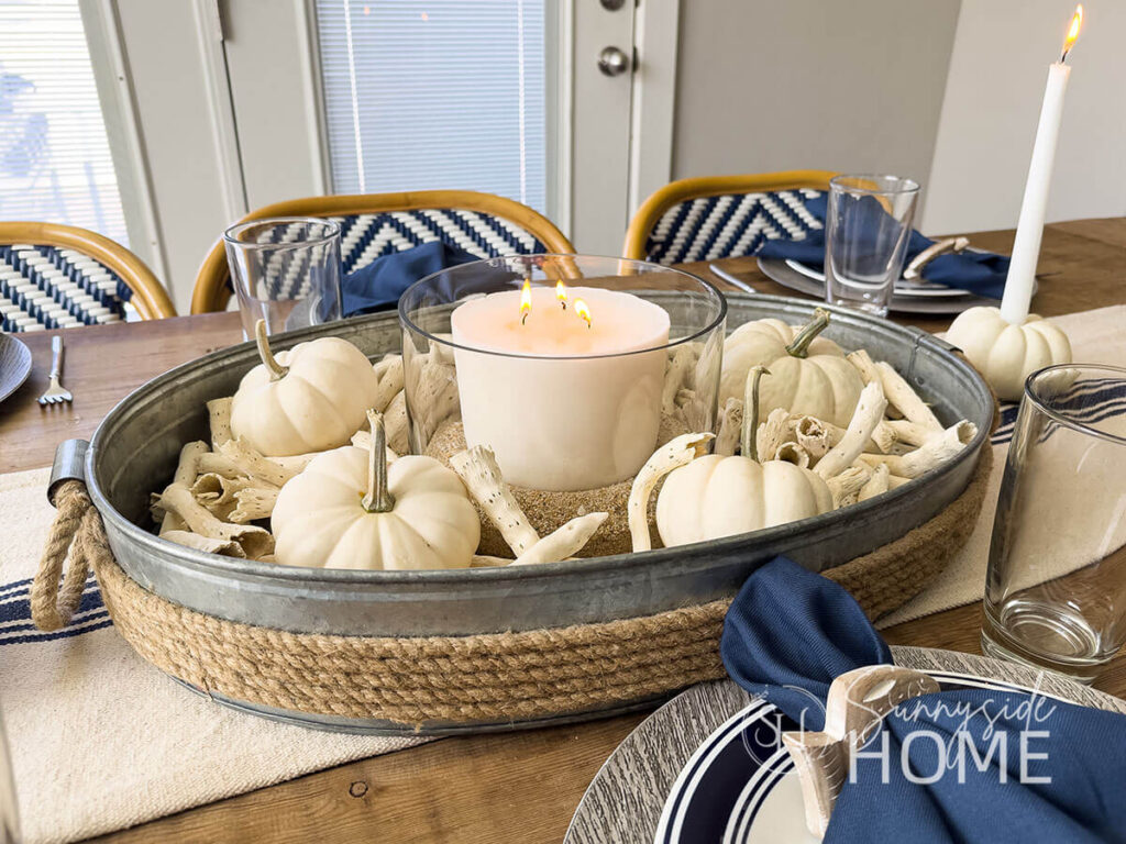 Coastal and fall Tablescape with a thrifted galvanized tray filled with drift wood, white mini pumpkins, and a white candle in a glass bowl with sand. The centerpiece is placed on a beige and navy striped table runner. The place settings feature a wood look dollar store charger, blue rimmed plate topped with a navy blue napkin in a wood fish napkin holder.