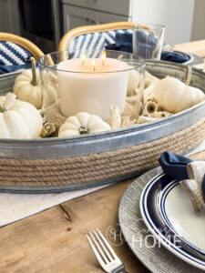 Coastal and fall Thanksgiving Table decor with a thrifted galvanized tray filled with drift wood, white mini pumpkins, and a white candle in a glass bowl with sand. The centerpiece is placed on a beige and navy striped table runner. The place settings feature a wood look dollar store charger, blue rimmed plate topped with a navy blue napkin in a wood fish napkin holder.