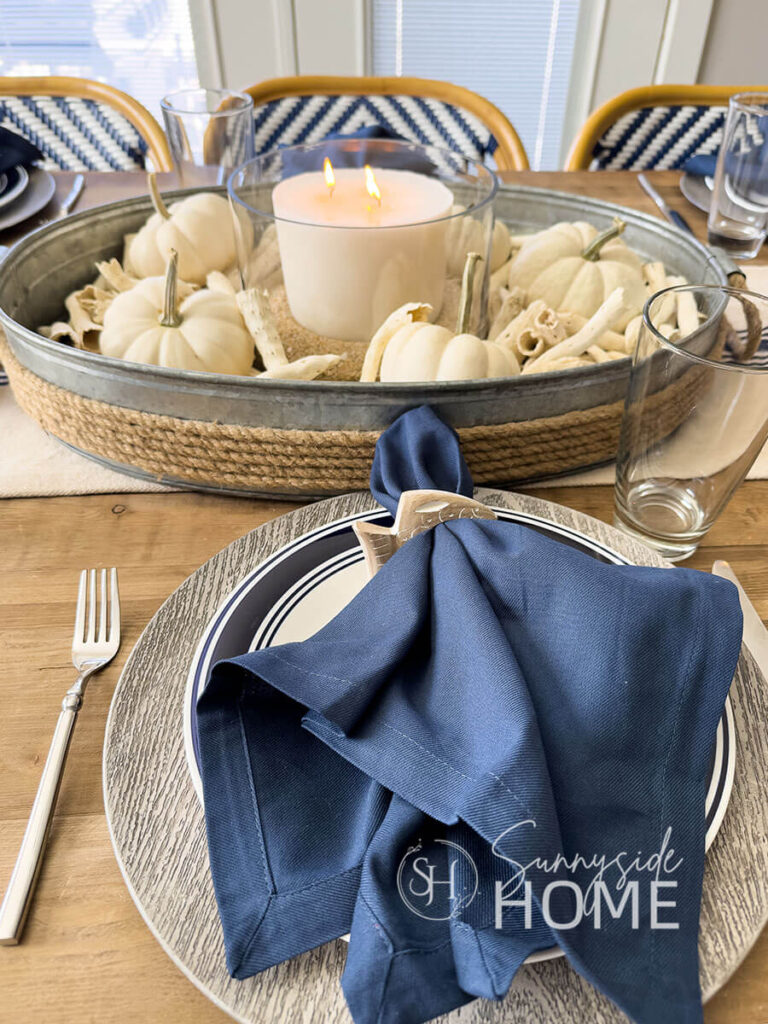 Coastal and fall Tablescape with a thrifted galvanized tray filled with drift wood, white mini pumpkins, and a white candle in a glass bowl with sand. The centerpiece is placed on a beige and navy striped table runner. The place settings feature a wood look dollar store charger, blue rimmed plate topped with a navy blue napkin in a wood fish napkin holder.
