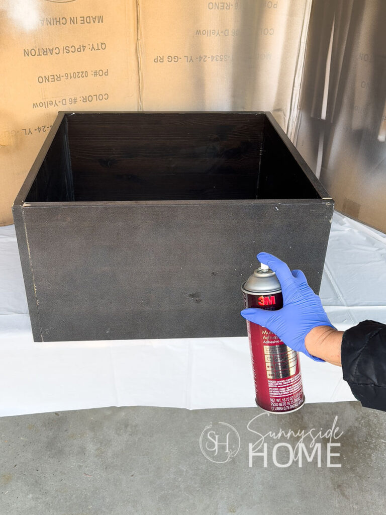 Woman sprays adhesive to a DIY tree collar built with scrap wood.