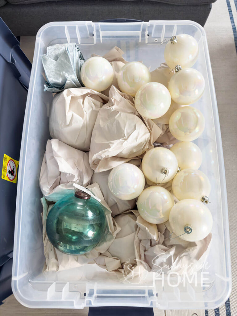 Large fragile glass ornaments are wrapped with paper and stored in a large clear storage bin.