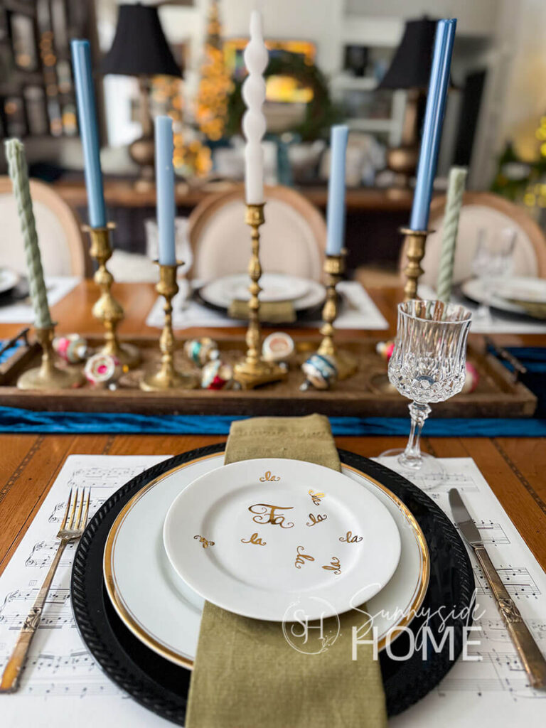 Dining table is set with thrifted gold rimmed dinner plates on a black charger with thrifted sheet music placemat. The dinner plate is topped with an olive green napkin and topped with a gold Christmas plate found at a yard sale.