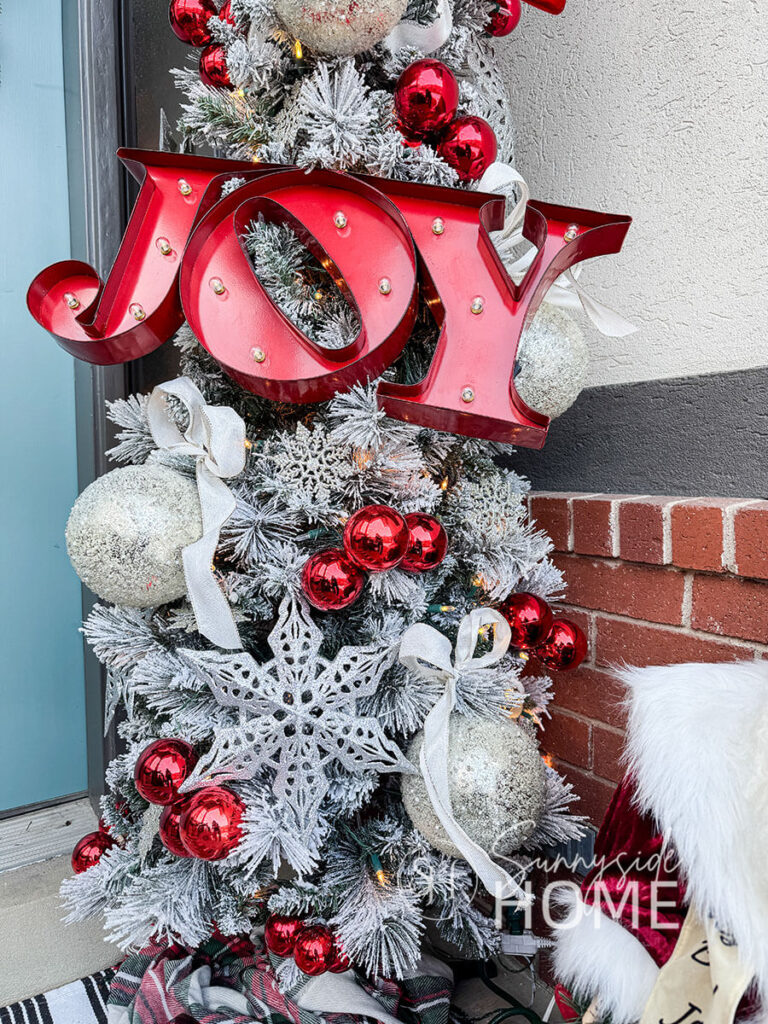 Close up view of decorated pencil tree on a small front porch with a large JOY marquee sign attaced the to the tree along with oversized icy ornaments, large bows, clustered red ball ornaments and large glittered snowflakes.