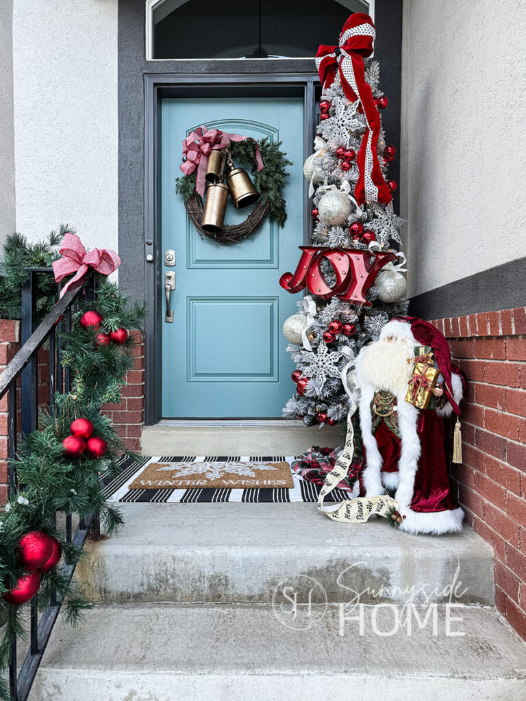 Decorate the front porch for Christmas with a pencil tree decorated with an oversized red velvet bow, oversized icy ornaments tied to the tree with ivory and gold ribbon, red ornament clusters and large glitter snowflakes. A vintage velvet Santa stands at the base of the tree and the railing is draped with a green garland embellished with clusters or red ball ornaments and red and white herringbone ribbon bows.
