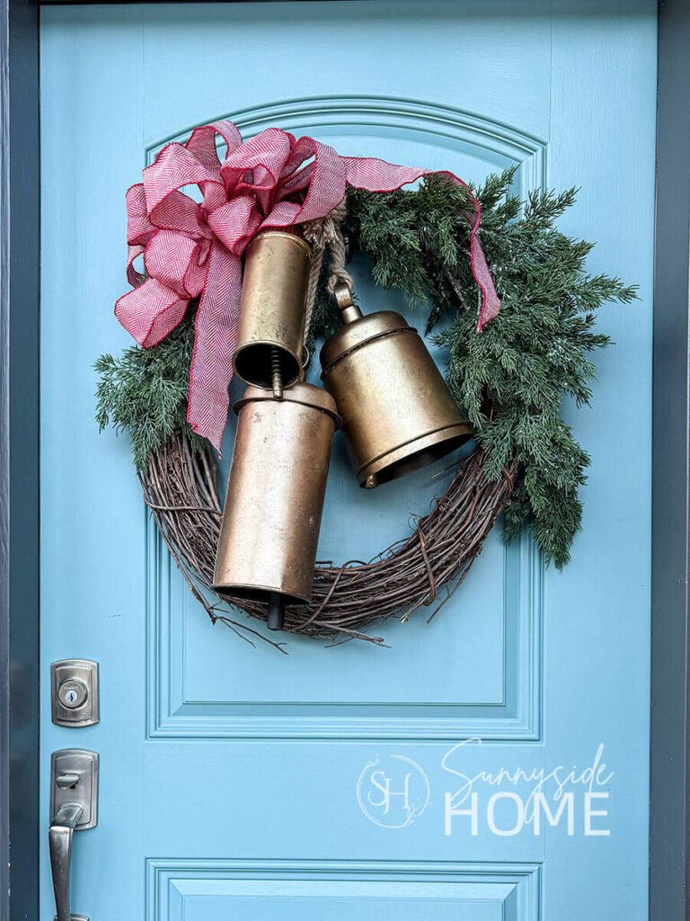 Christmas wreath with cedar greens, three large faux brass bells and a red and white herringbone patterned bow.