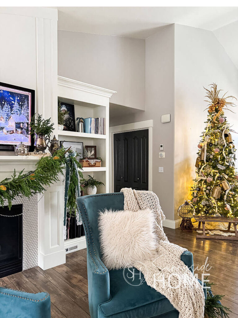 Family room is decorated for Christmas with a cuddly throw blanket and faux fur pillows, garland on the mantle, and in the background is the decorated entryway Christmas tree.