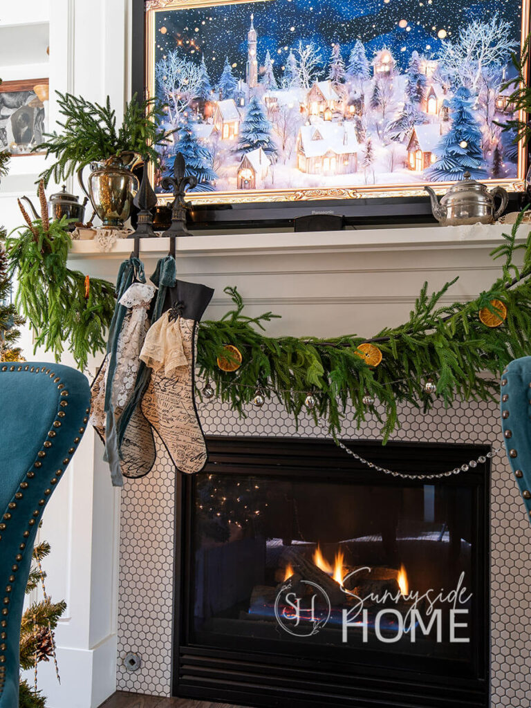 Fireplace mantle decorated for Christmas with a Norfolk pine garland, embellished with dried orange slices, chandelier crystals and mercury glass ornaments. Silverplated pieces with pine stems and doilies decorate the mantle.
