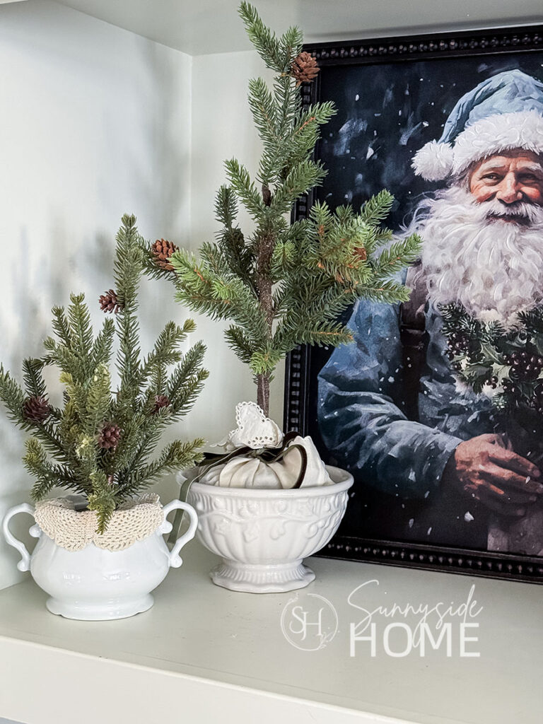 Pine trees are wrapped with vintage linens and tied with velvet ribbon and placed in thrifted footed bowls, framed Santa painting in the back of the shelf.