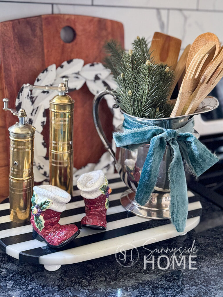 Greenery is added to a silverplated pitcher that holds cooking utensils and tied a blue velvet ribbon, Santa boot salt and pepper shakers styled on a marble tray.