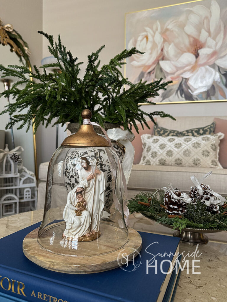 Christmas decor on the coffee table, cloche with a nativity, silver footed bowl filled with cedar greens, and frosted pinecones, vase filled with norfolk pine stems and tied with a ivory and gold bow.