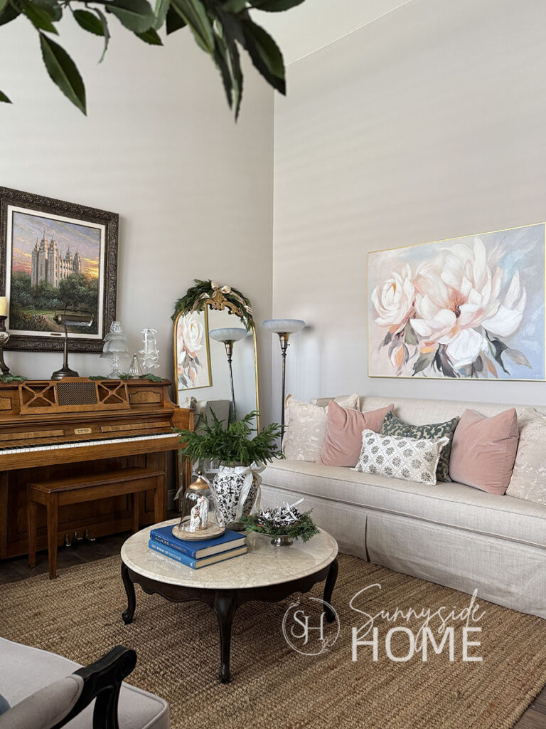 Living room is decorated for Christmas with cedar swags on the floor mirror, embellished with a gold and ivory ribbon, beaded snowflake pillow on the sofa, coffee table styled with Norfolk pine stems in vase, nativity under a cloche and silver bowl with cedar stems and frosted pinecones.
