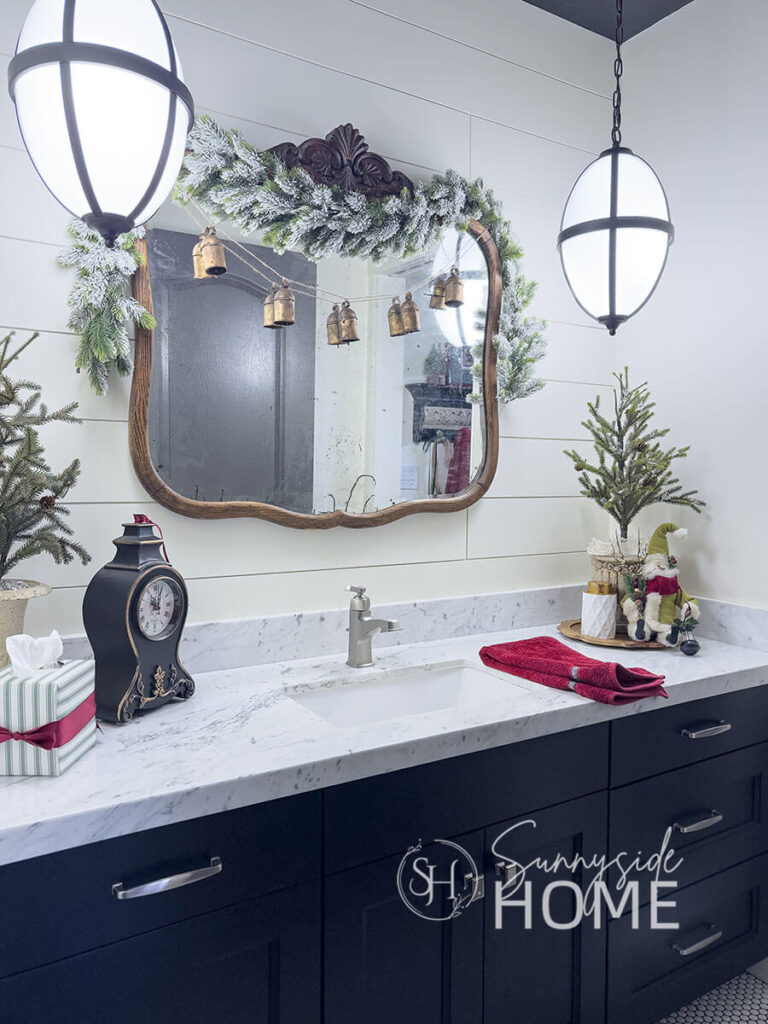 Over the bathroom mirror is draped a flocked garland embellished with DIY faux brass bells, and the vanity is decorated for Christmas with small pine trees in thrifted pots.