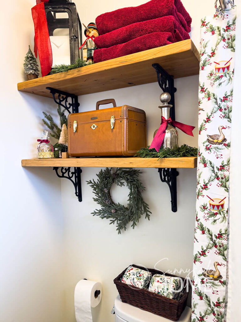 Bathroom shelves are decorated for Christmas with stack red towlels, an old lantern filled with rolls of toilet paper, a nutcracker, mini trees and wreaths.