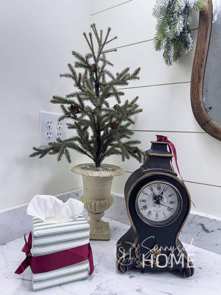 Pine tree in a vintage thrifted pot, thrifted black clock embellished with a burgundy ribbon and a tissue box is covered in gift wrapping paper and embellished with a burgundy ribbon in the bathroom.
