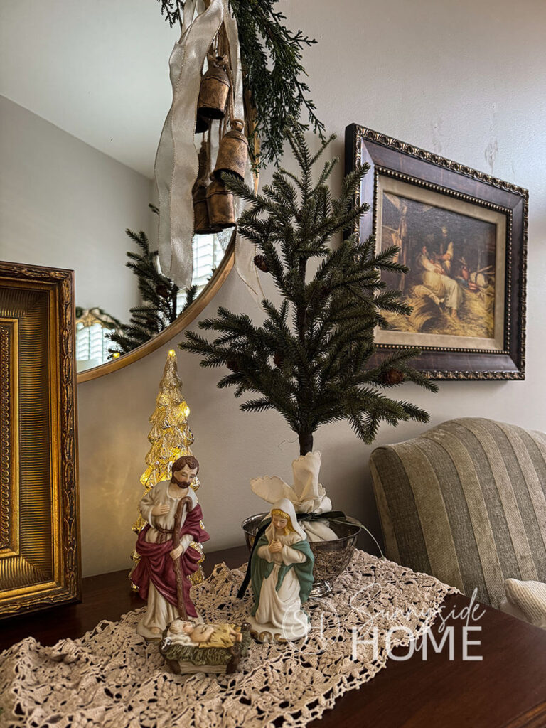 Grandma's nativity set styled on the antique dresser on a vintage crochet table runner. A painting of the nativity is hung on the wall and the round mirror is embellished with a cedar garland, faux DIY brass bells and ivory and gold ribbon.