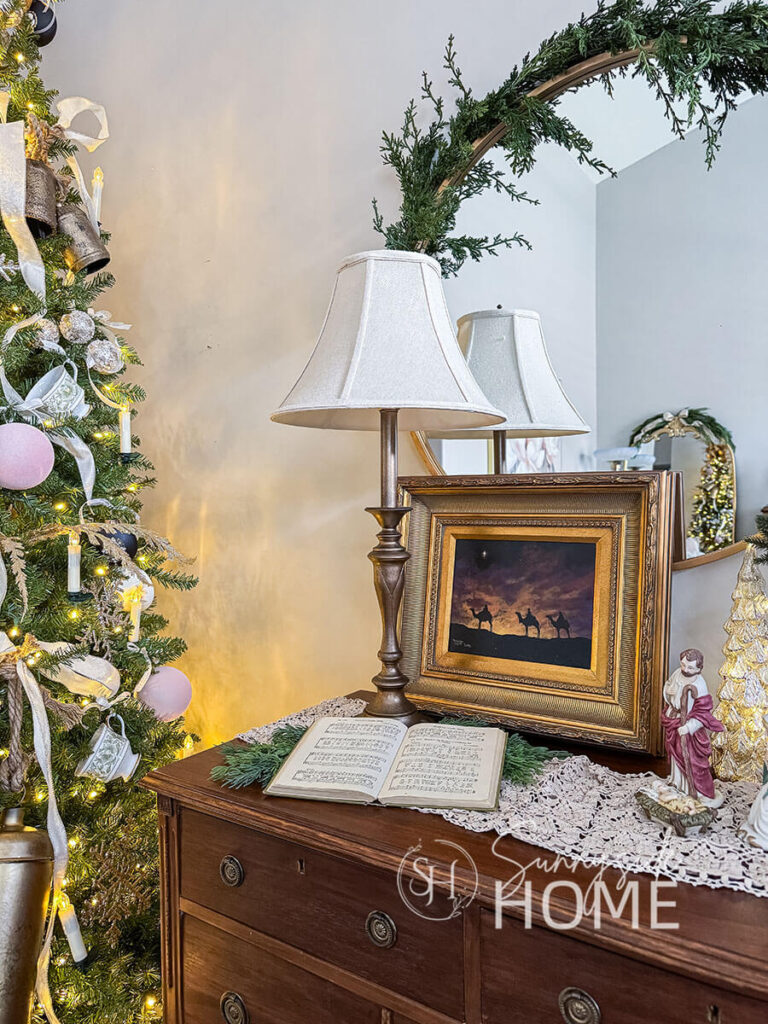 An antique dresser serves as the entryway table and is decorated with a faux antique brass lamp, Christmas wisemen art, old hymnal, greens, and grandma's nativity set. Reflecting in the mirror you can see the lighted Christmas tree, with a garland embellishing the mirror.