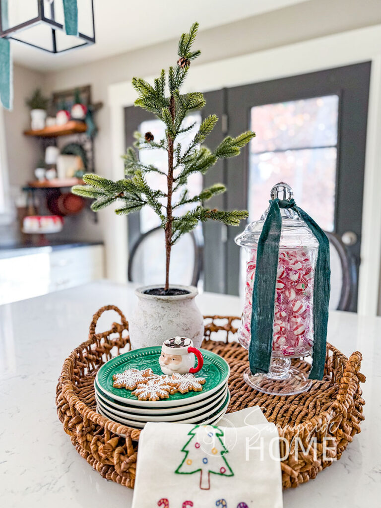 In the kitchen the island is decorated for Christmas with a pine tree in a pot, apothecary jar filled with peppermints and embellished with blue velvet ribbon and a stack of green plates with faux gingerbread cookies and a Santa mug of hot cocoa styled in a wicker tray.