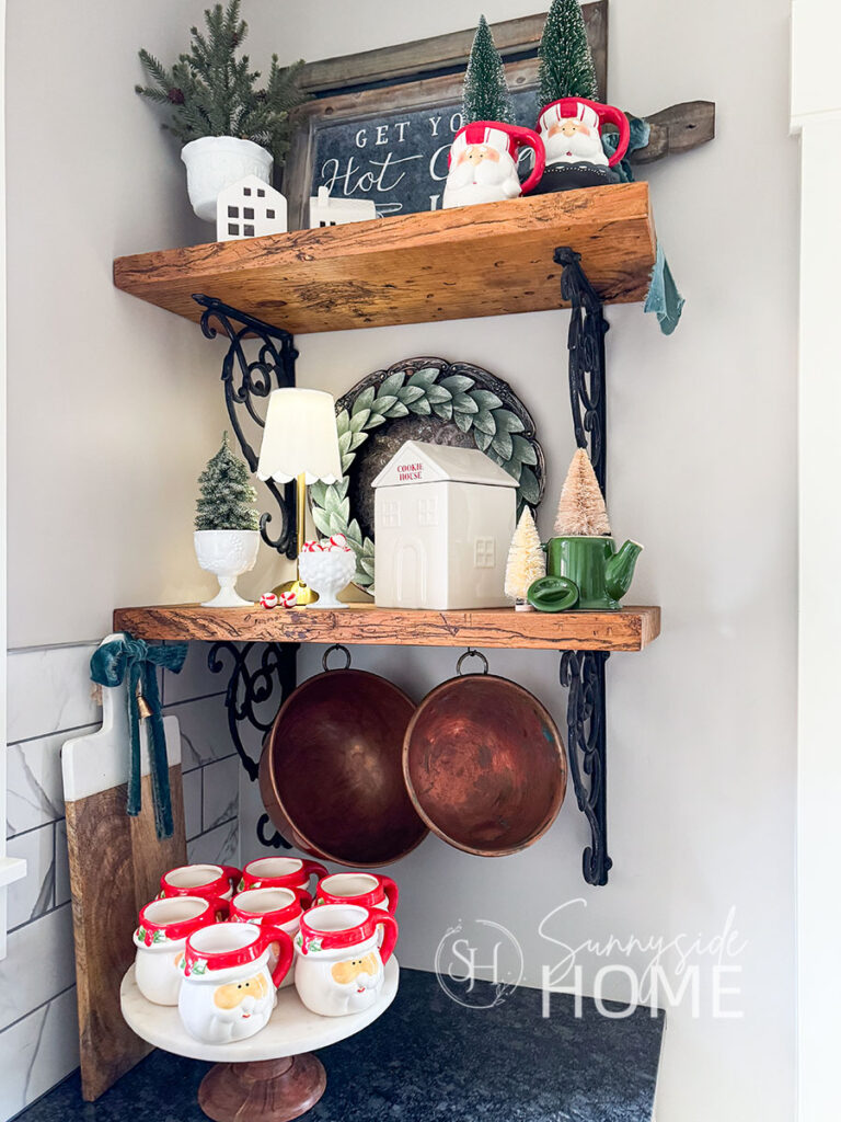 Open shelves in kitchen are decorated for Christmas with sisal trees in Santa mugs, mini trees in milk glass footed bowls, hot cocoa sign, Christmas cookie house, and Santa mugs on a pedestal tray.
