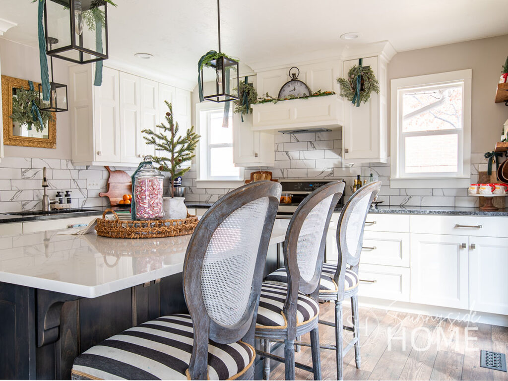 Kitchen decorated for Christmas with cedar greenery on pendent lights tied with blue velvet ribbon, wreath with blue velvet ribbon hung on cabinet doors, and a wicker tray styled with an apothecary jar filled with peppermints, Christmas tea towel and a potted mini pine tree.