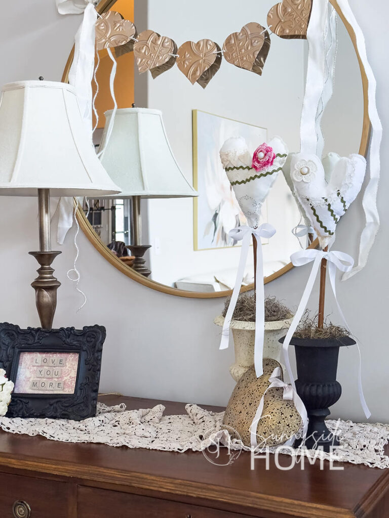 DIY Valentine heart decor styled on an entry table with faux vintage tile heart garland, brassed heart, black framed "love you more" decor and a faux antique brass lamp.