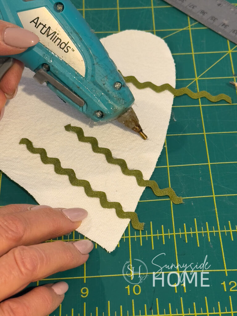Woman applies a narrow bead of hot glue to linen heart to apply a strip of olive green ric rac trim.