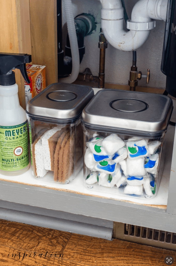 Dishwasher cleaning tabs and sponges are decanted into clear storage containers under the kitchen sink.