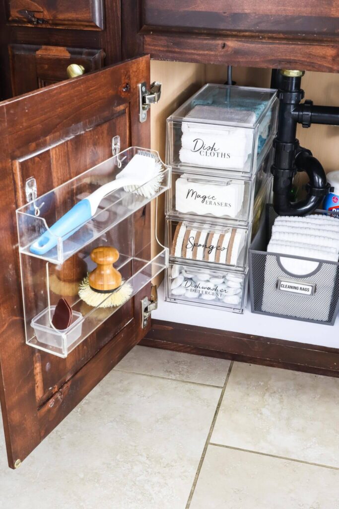 Clear spice rack is mounted to the cabinet door under the sink with hooks to store cleaning brushes.