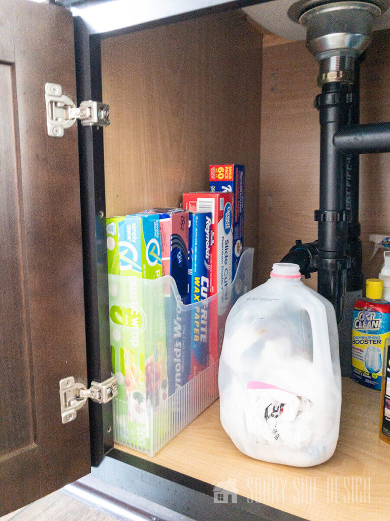 A magazine holder is used to organize kitchen wraps under the kitchen sink along with an old milk jug for grocery bags.