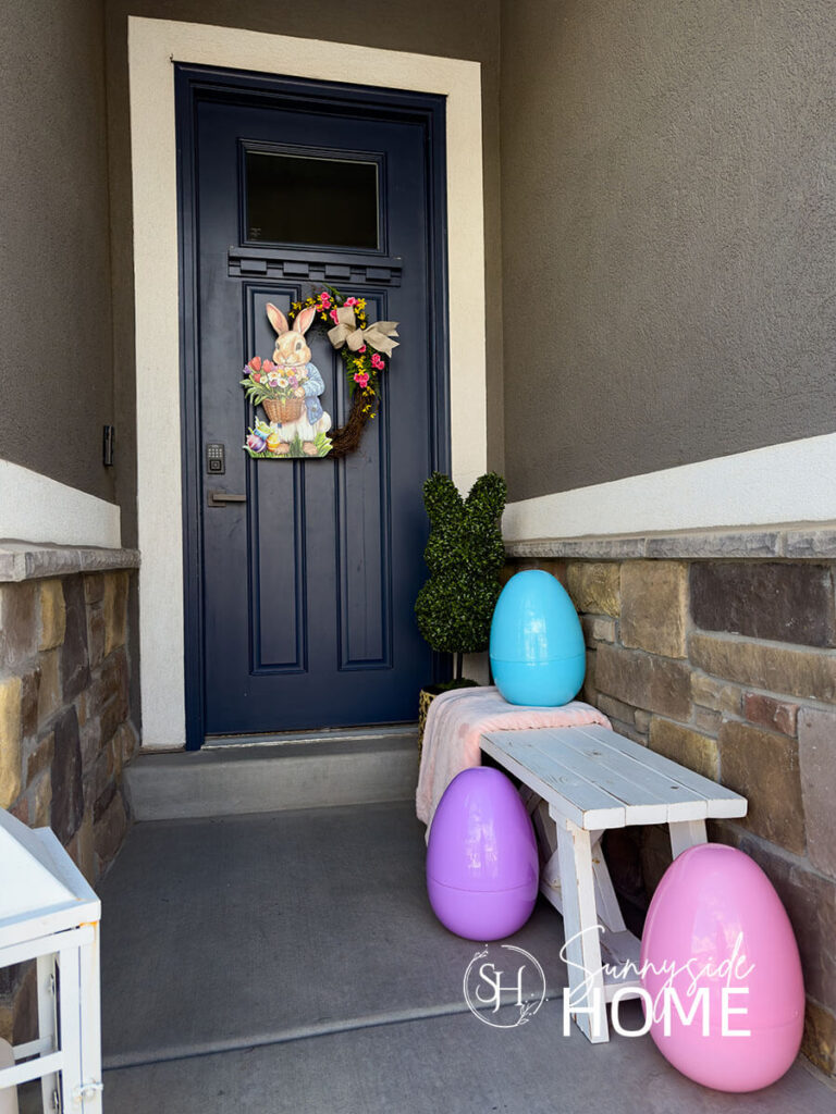 Styled DIY Easter wreath with a bunny topiary and large pastel Easter eggs on a small front porch.