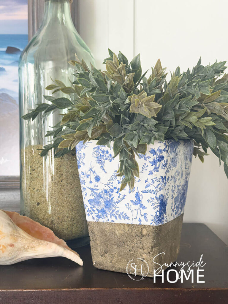 Blue and white decoupaged cement vase styled with greenery, conch shell and a jug of sand.