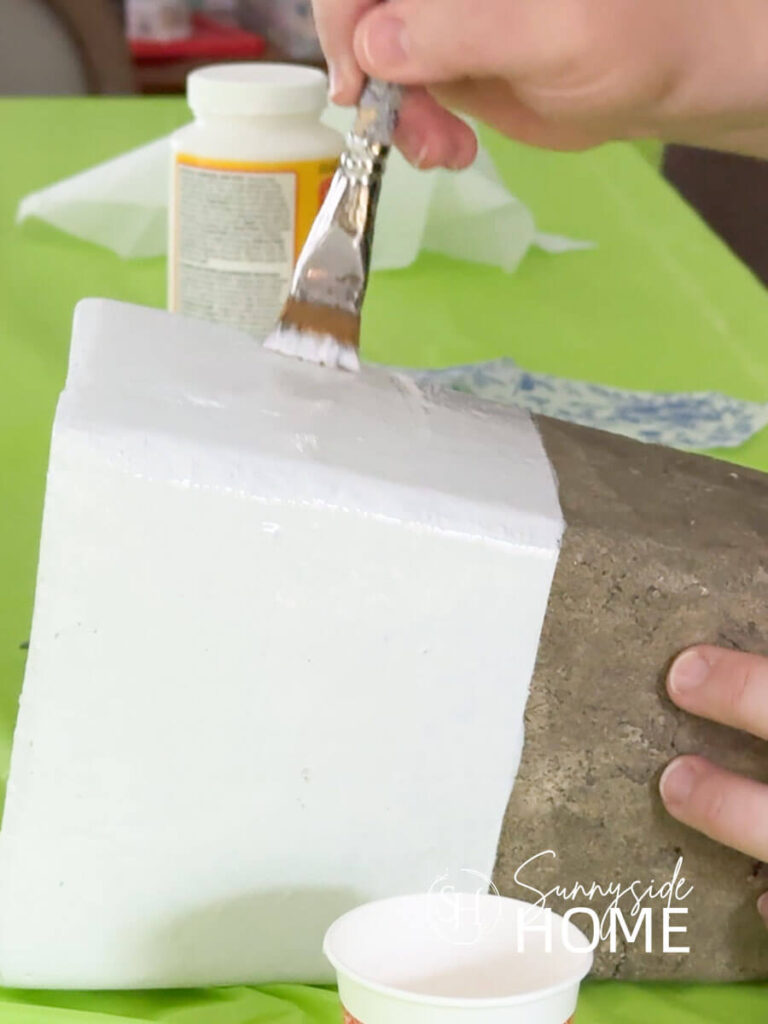 A woman applies Mod Podge to a painted section of a cement vase.