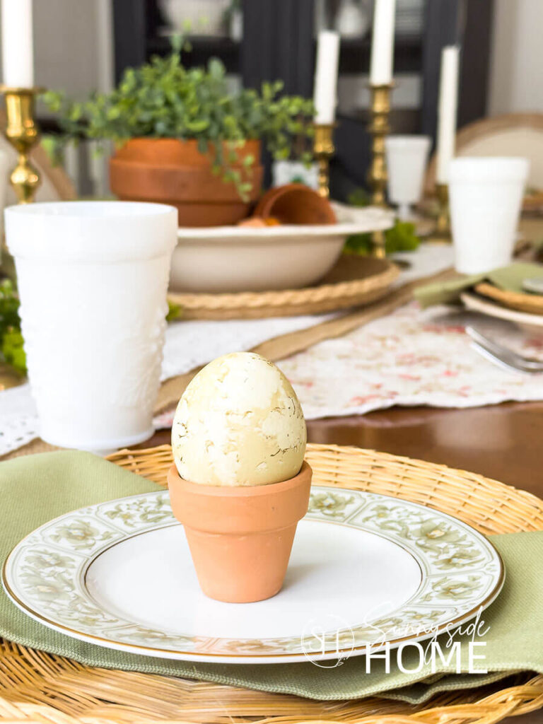The Easter tablescape is placed on burlap and lace table runners and topped with a large white bowl filled with stacked and toppled clay pots and greenery, brass candlesticks, . Easter place settings, off white plates are placed on rose bud fabric and topped with a wicker plate holder, green floral rimmed bread place and a mini clay pot holds a green dyed egg.