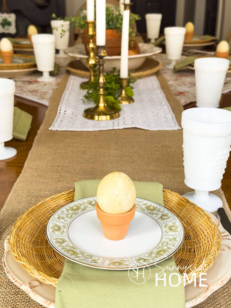 The Easter tablescape is placed on burlap and lace table runners and topped with a large white bowl filled with stacked and toppled clay pots and greenery, brass candlesticks, . Easter place settings, off white plates are placed on rose bud fabric and topped with a wicker plate holder, green floral rimmed bread place and a mini clay pot holds a green dyed egg.