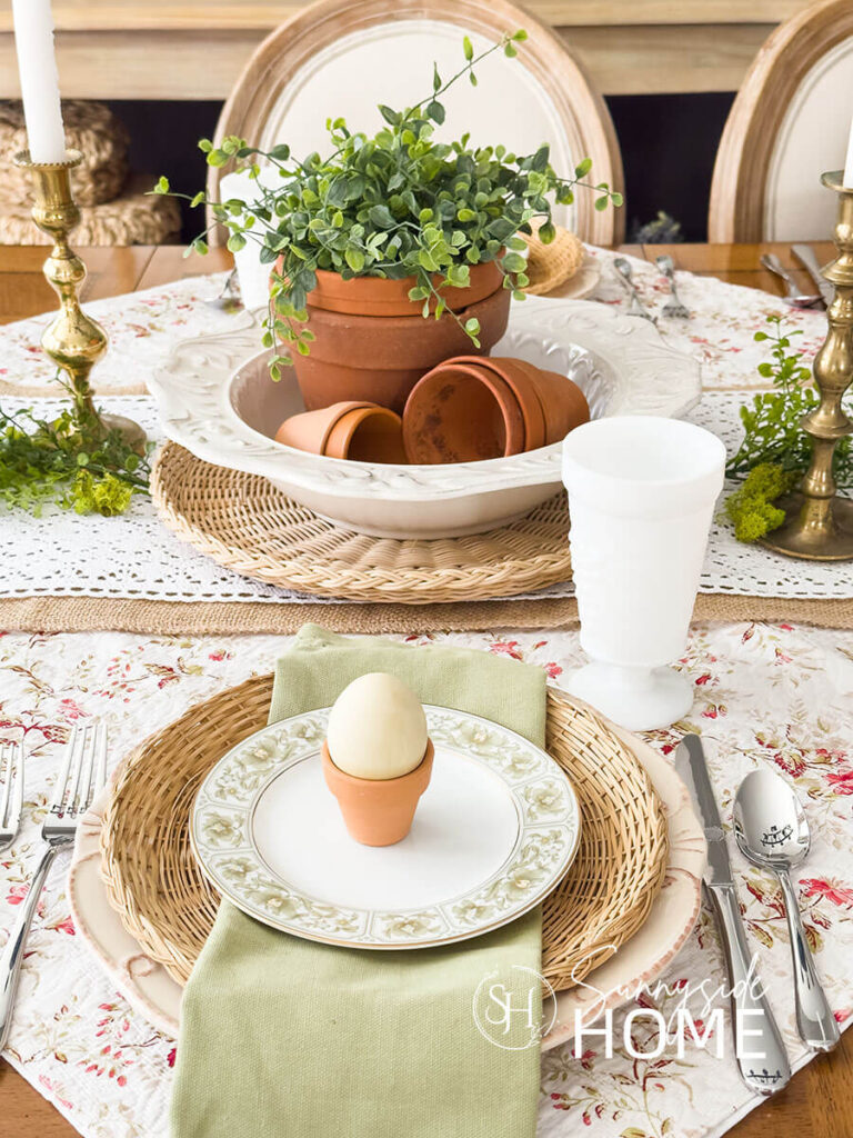 The Easter centerpiece is placed on burlap and lace table runners and topped with a large white bowl filled with stacked and toppled clay pots and greenery. Easter place settings, off white plates are placed on rose bud fabric and topped with a wicker plate holder, green floral rimmed bread place and a mini clay pot holds a green dyed egg.