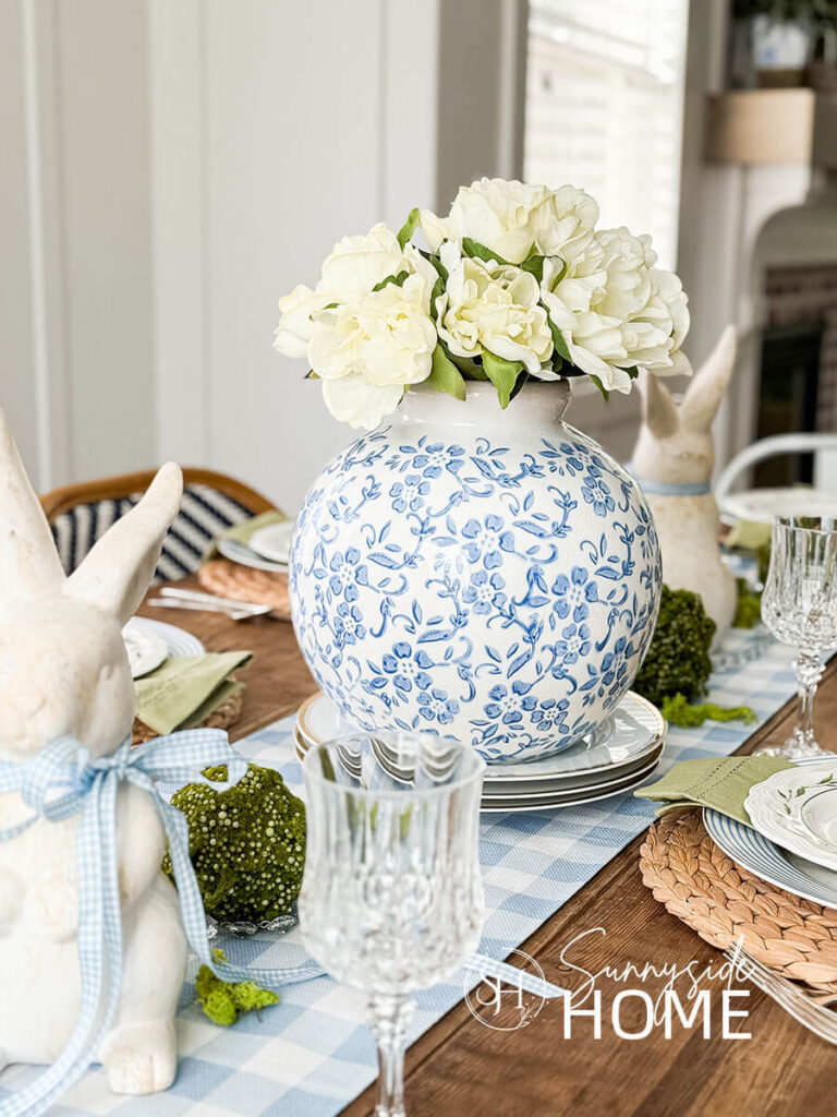 Blue and white Easter table settings with a blue and white vase with peonies centerpiece on a blue and white gingham table runner embellished with bunnies and moss balls.