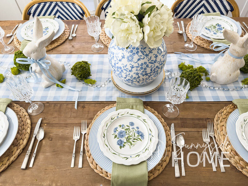 Blue and white Easter tablescape with a blue and white vase with peonies centerpiece on a blue and white gingham table runner embellished with bunnies and moss balls, water hyacinth placemats, blue and white striped plate, sage green napkin and a floral plate.