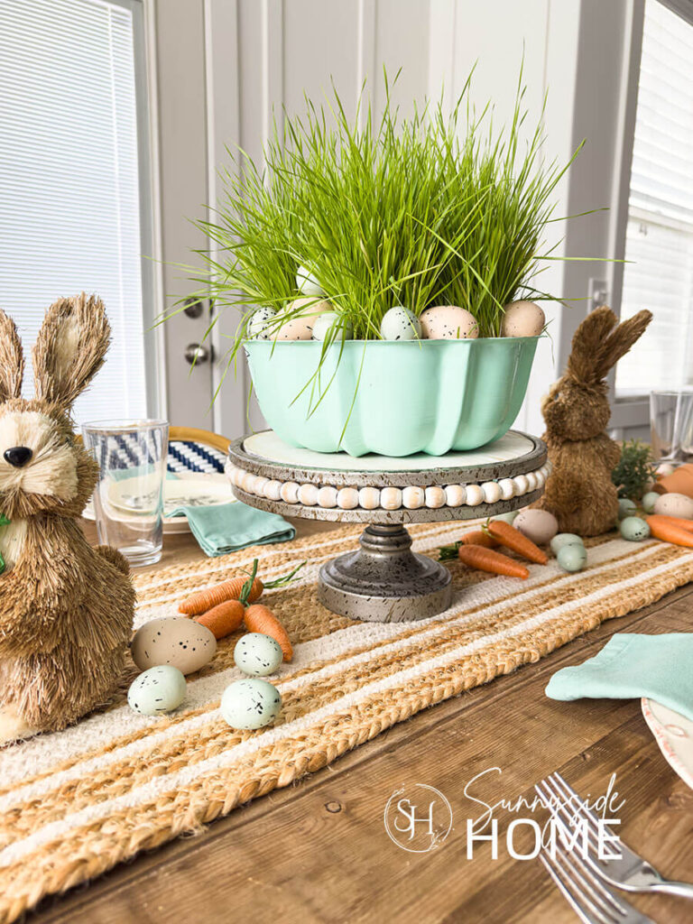 Easter table centerpiece starts a vintage aqua bundt pan placed on a cake stand and is filled with wheat grass and Easter eggs, clay pots, scattered string carrots, greenery eggs and bunnies are placed on a jute table runner.