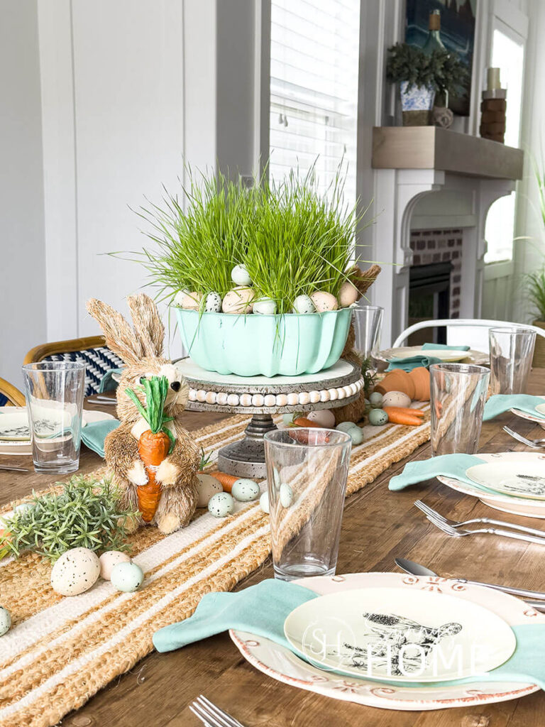 Easter tablescape with off white dinner plate topped with an aqua napkin and a disposable plastic bunny plate. The centerpiece a vintage aqua bundt pan is placed on a cake stand and is filled with wheat grass and Easter eggs, clay pots, scattered string carrots, greenery eggs and bunnies are placed on a jute table runner.