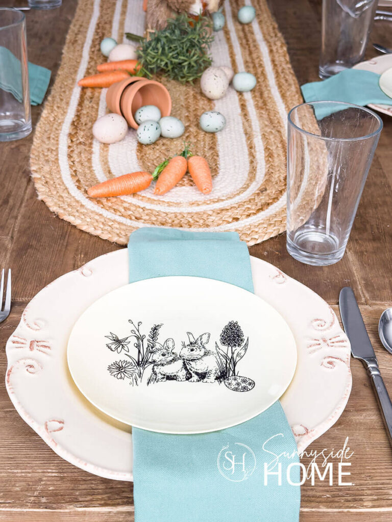Easter table settings, Off white dinner plate topped with an aqua napkin and a disposable plastic bunny plate. The centerpiece of clay pots, scattered string carrots, greenery and eggs are placed on a jute table runner.