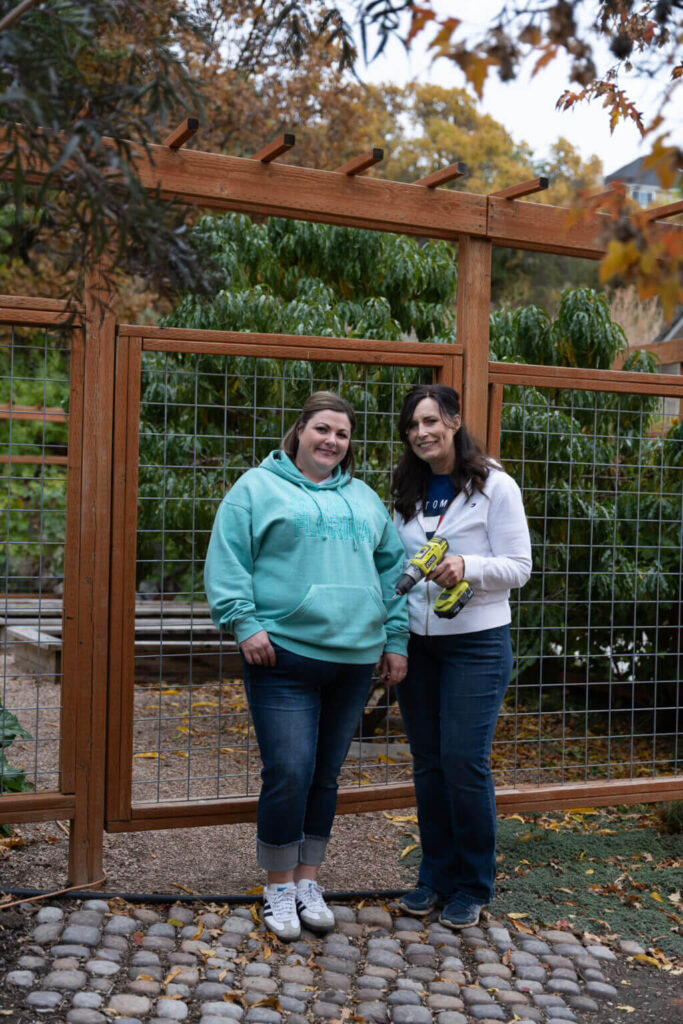 Two women stand outside the gate for this 8' tall garden enclosure, one woman holds a drill.