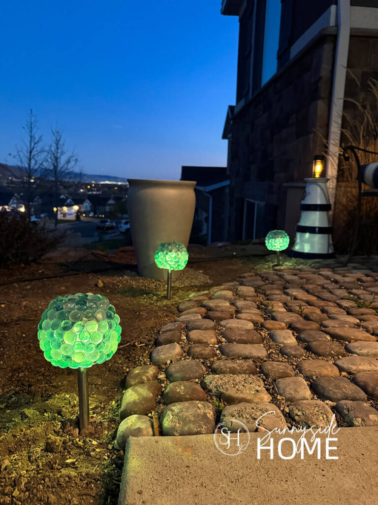 Three solar orbs along the edge of a cobblestone path leading to a garden bench. The orbs are made with dollar store solar lights.