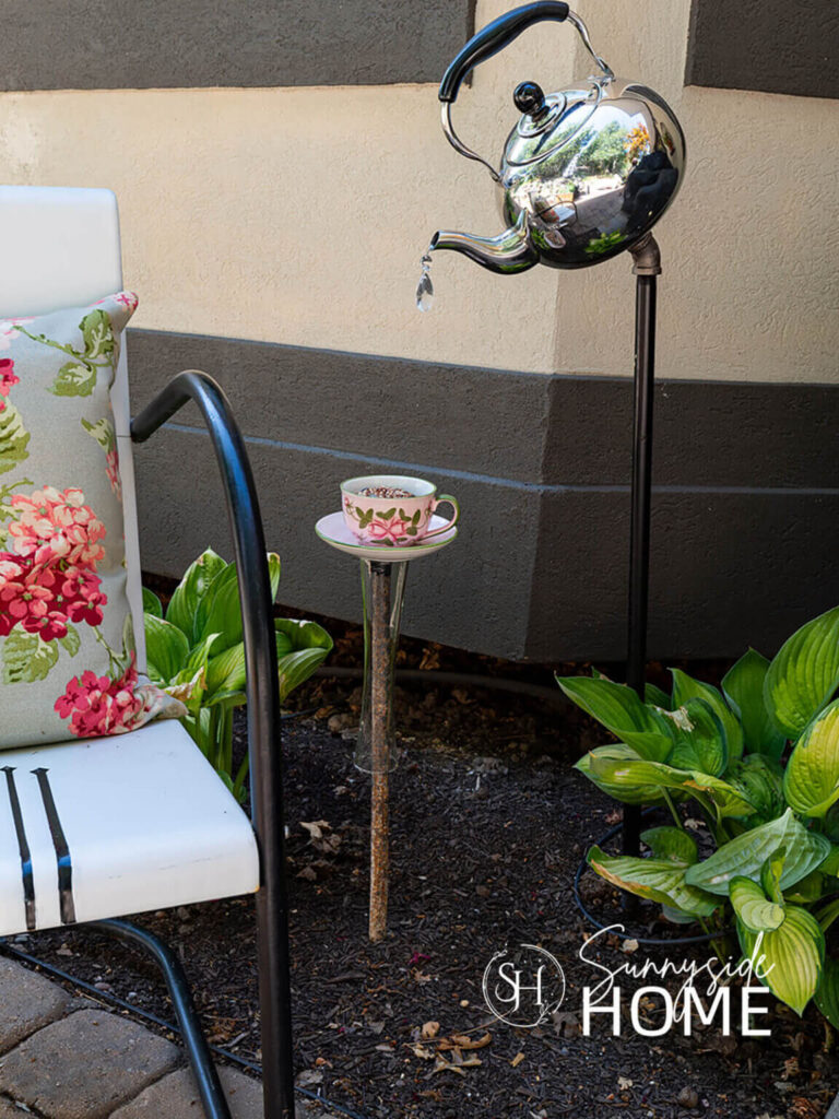 Whimsical teapot and cup as fun garden decor alongside a vintage metal chair with a floral pillow.