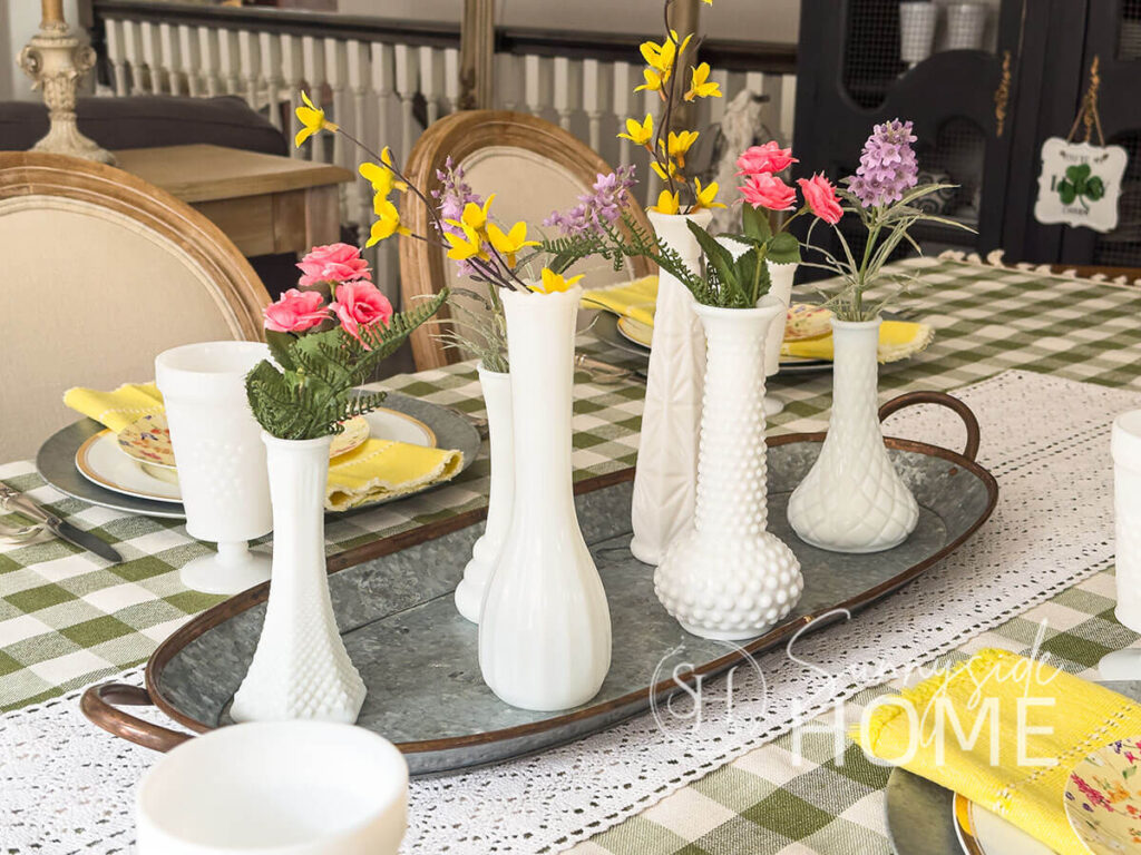 Easter centerpiece, a galvanized tray topped with 6 milk glass bud vases with forsythia, rose buds and lavender with a olive green gingham tablecloth, crochet lace table runner.