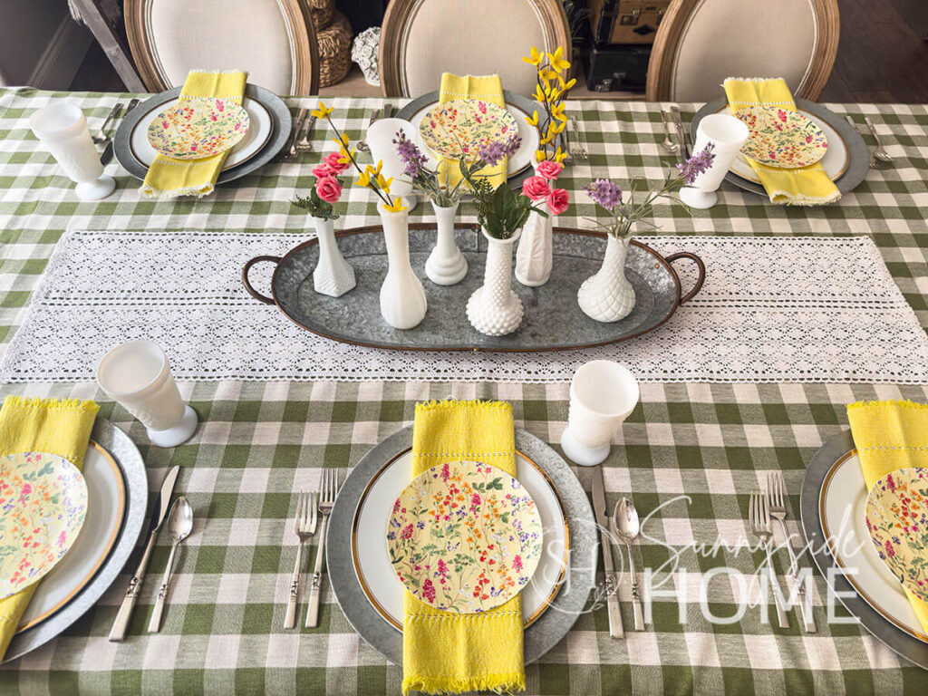 Easter tablescape with a olive green gingham tablecloth, crochet lace table runner, topped with a galvanized tray. In the tray are 6 milk glass bud vases with forsythia, rose buds and lavender. At each table settings is a galvanized charger, topped with a gold rimmed plate, yellow hemstitched napkin and then topped with a disposable floral plastic plate. Vintage milk glass goblets and silverware.