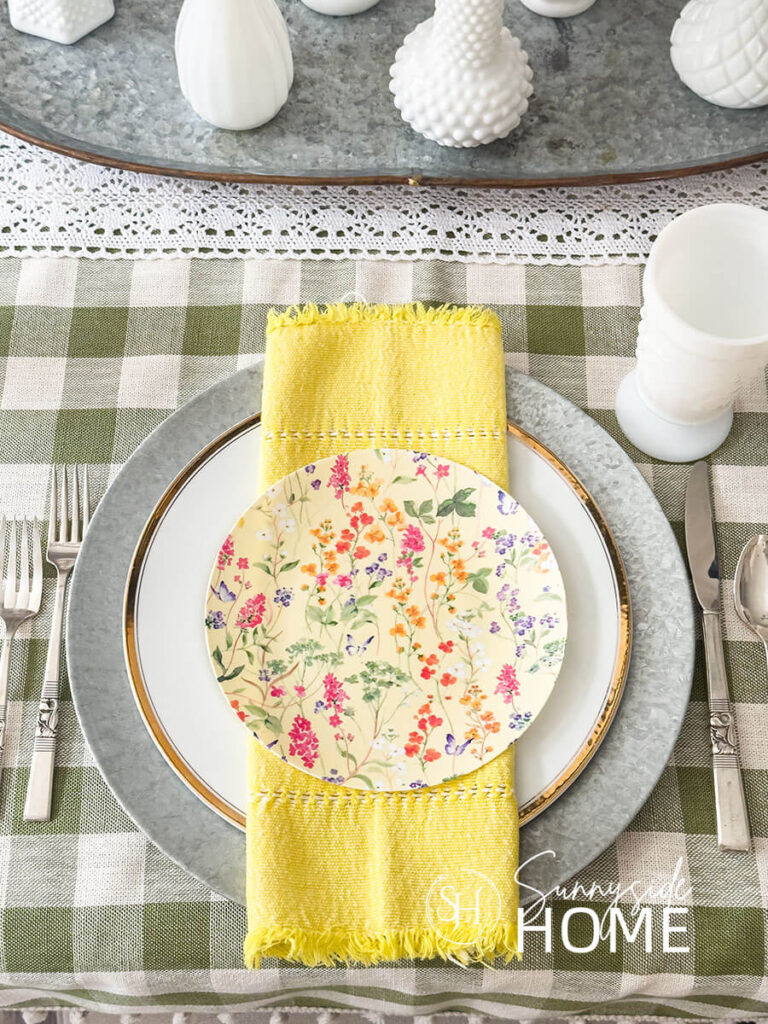Simple Easter table settings with a galvanized charger on a olive green tablecloth, topped with a gold rimmed plate, yellow hemstitched napkin and topped with a disposable floral plastic plate.