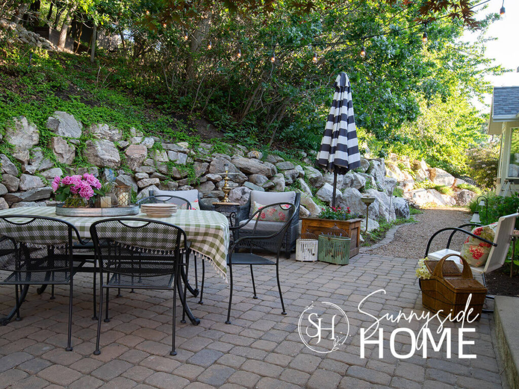 Backyard ideas include a styled patio with a dining table with a green gingham tablecloth, peonies in a vase, tray with lanterns, stacked plates, floral pillows on patio chairs, planter box with dahlias, bird cages, picnic basket filled with hydrangeas and string lights overhead.